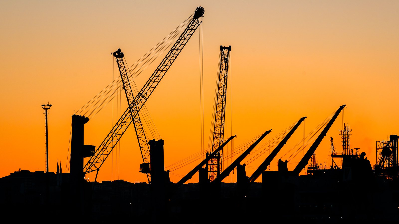 Cargo cranes and a docked vessel in silhouette against an orange sky.