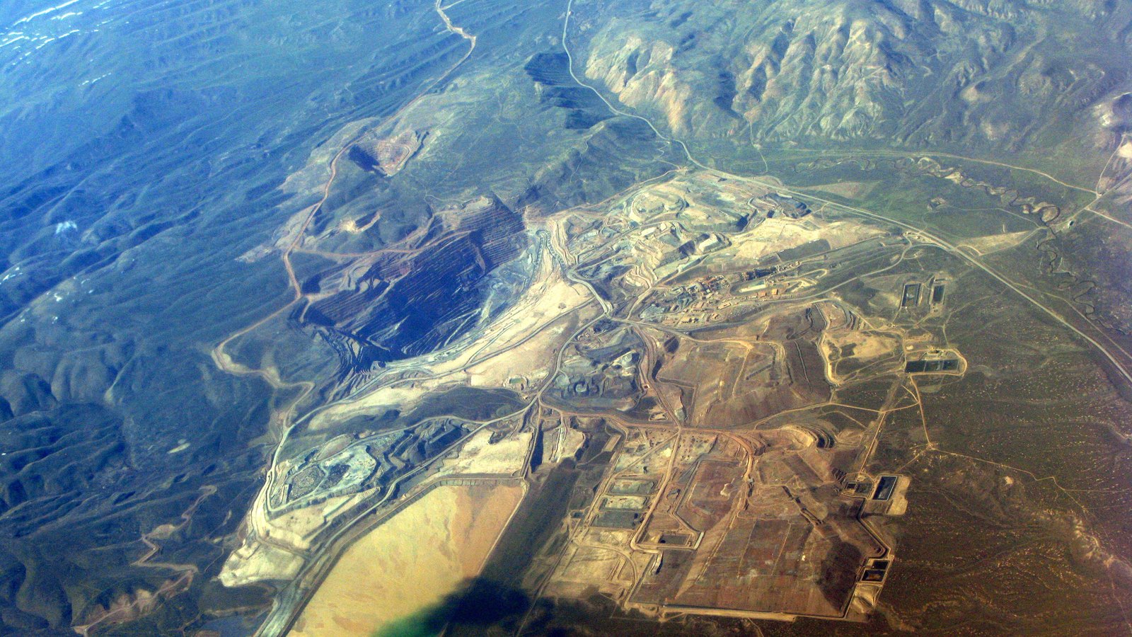 An aerial view of the Gold Quarry open-pit mine outside Carlin, Nevada, with terraced pits, pale waste rock, a broad flat basin and roads running across brown high desert.