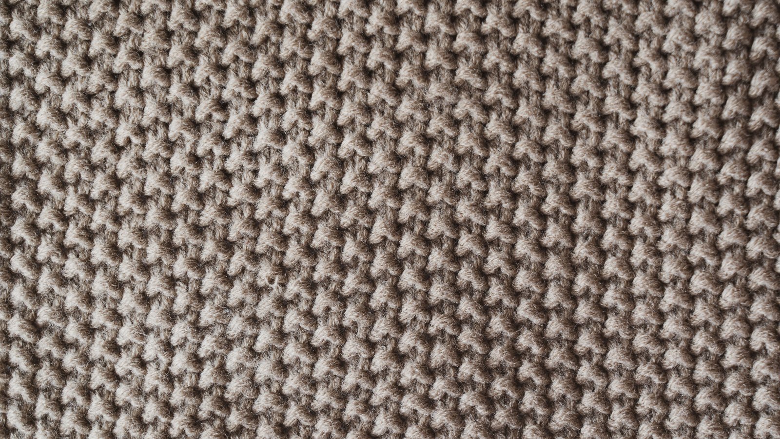 An extreme close photograph of knitted woollen cloth in a plain greyish brown, showing regular rows of small raised stitches repeating evenly across the entire frame, with individual fibres visible on each loop and no seam, edge or label in view.