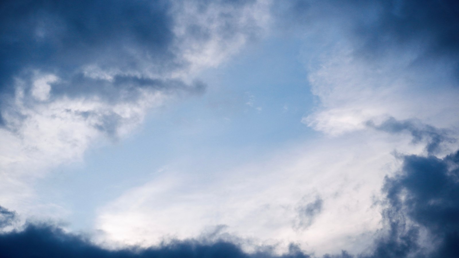 A break of pale blue sky ringed by heavy grey and white cloud, dark banks closing in along the bottom edge and the upper left corner, with thin bright cloud feathering across the open centre. The frame holds sky only. No horizon, land, water, people, buildings, signage or text are in view.