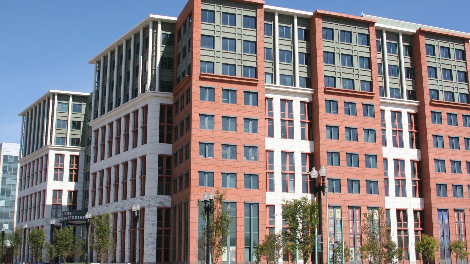 The United States Department of Transportation headquarters in Washington, a long facade of red and white masonry with ranks of square windows, street lamps and young trees along the pavement, and the building's carved name plate partly visible at street level.