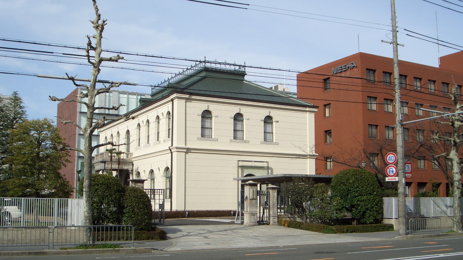 A two storey cream coloured building with arched windows and a dark green hipped roof stands behind a low hedge and a gate, with a red brick office block behind it at the right carrying the word NISSHA in metal letters near the roofline. Bare street trees, overhead cables and a small road sign frame the view and the pavement in front is empty. No people or vehicles are in view.