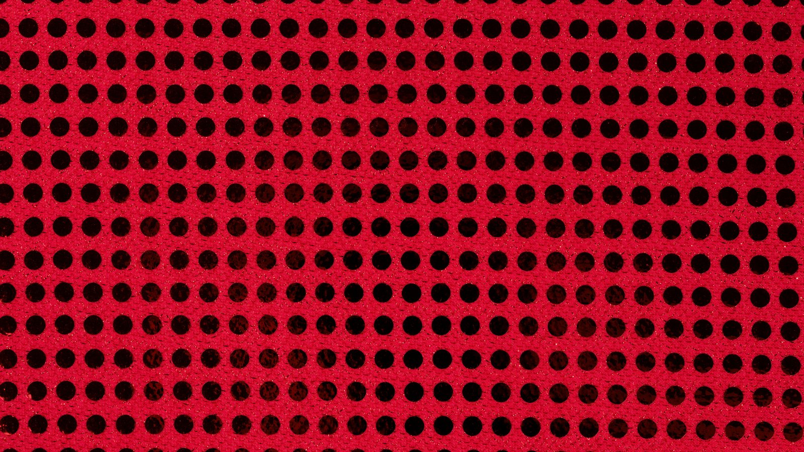 A close photograph of a flat red textured surface filling the whole frame, pierced by straight rows of evenly spaced round black holes running edge to edge, with the red material between the holes flecked by tiny lighter specks.
