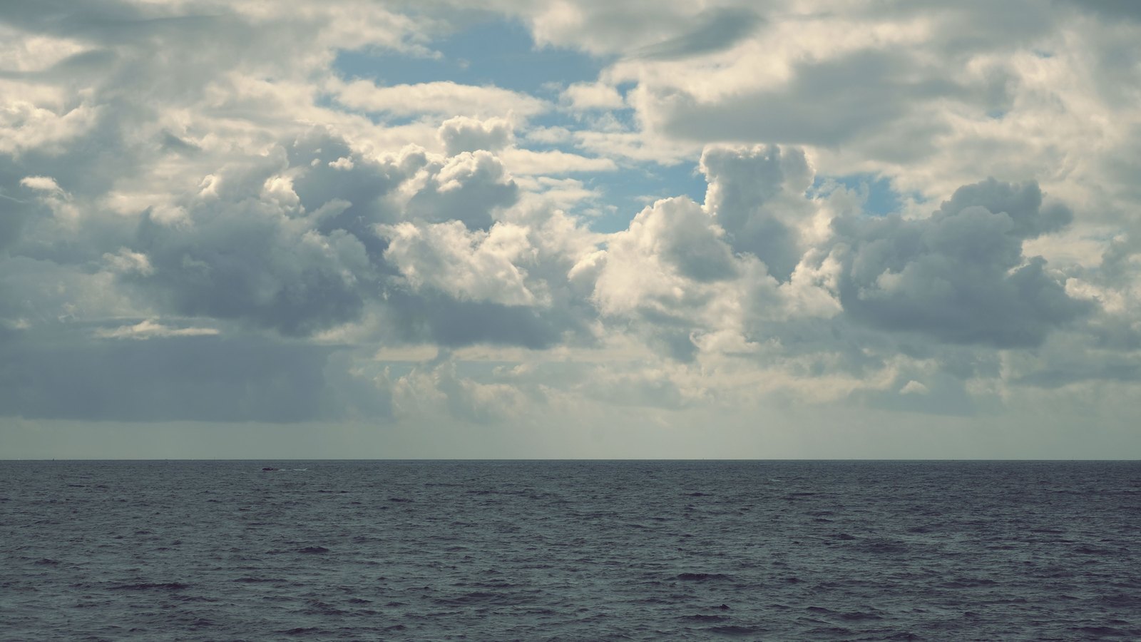 Open grey blue ocean seen from sea level under a broken deck of white cumulus cloud, with an empty horizon and no land in sight.