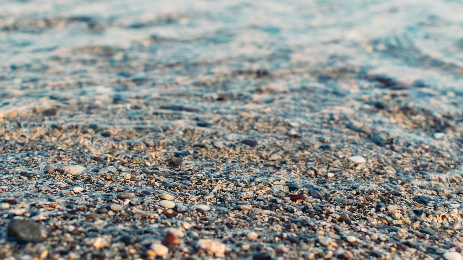 A low close view of a bed of small rounded pebbles and coarse grit in browns, greys and blues, the nearest stones sharp and the further ones softening under a thin sheet of clear water. No text, brand mark, person, vessel or shoreline is in view.