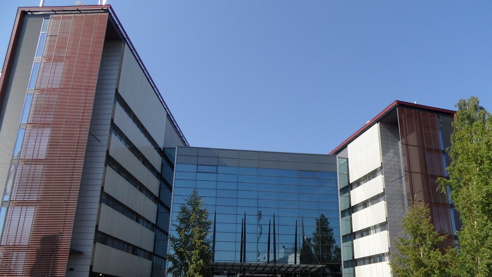 Nokia's head office in Espoo, Finland: two office wings clad in rust-red louvres and pale panels flank a glass atrium that reflects birch trees and a row of slim masts under a clear blue sky.