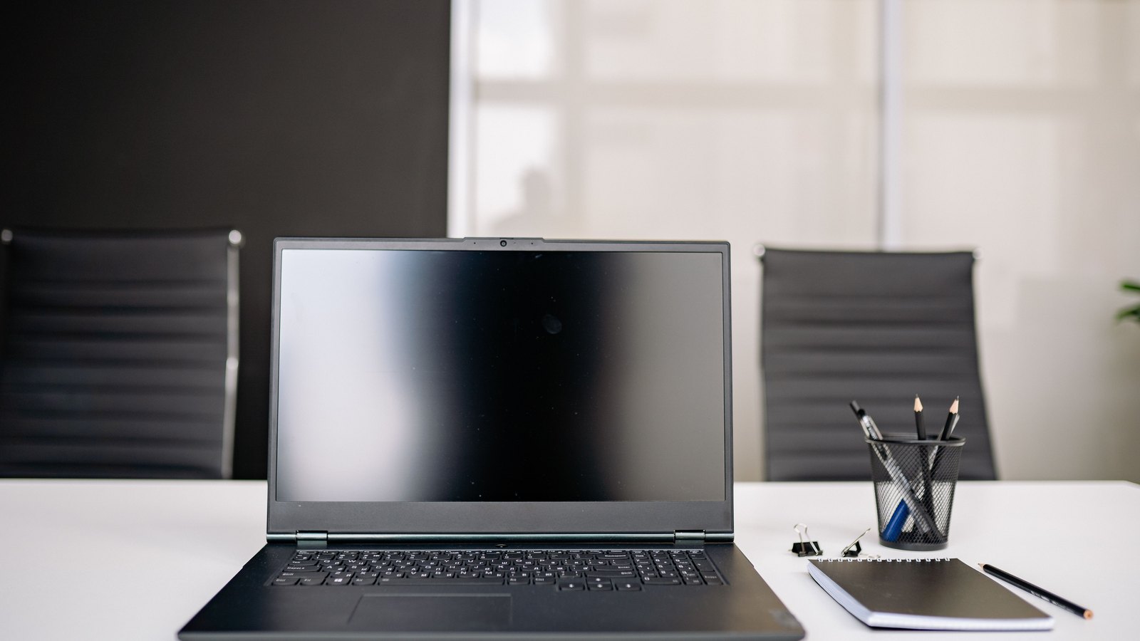 An open laptop with a completely black switched off screen standing on a white table, two empty office chairs behind it, and a wire pen pot and a closed notebook to the right.