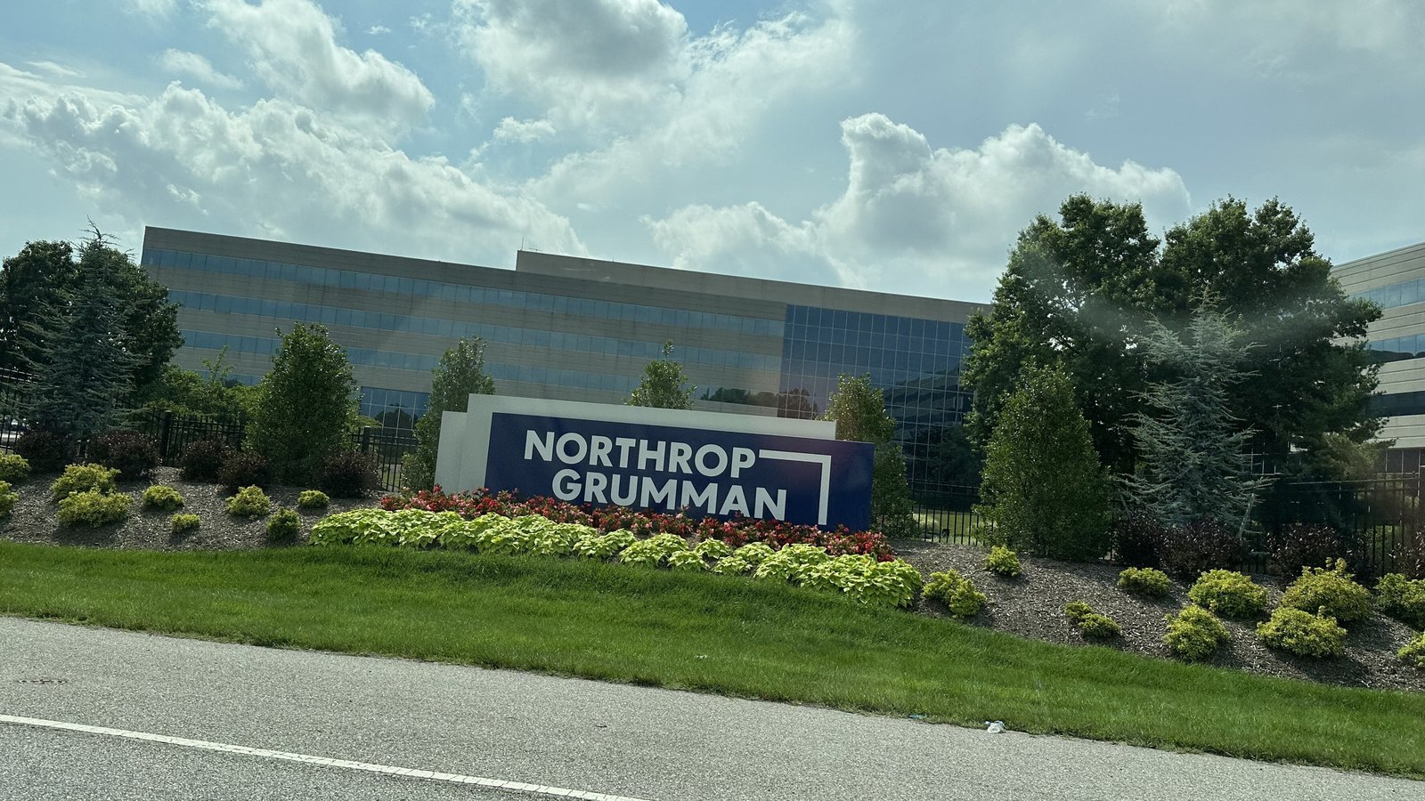 The Northrop Grumman name on a blue monument sign in a landscaped bed in front of the company's Mission Systems office building in Linthicum, Maryland, under a cloudy sky.