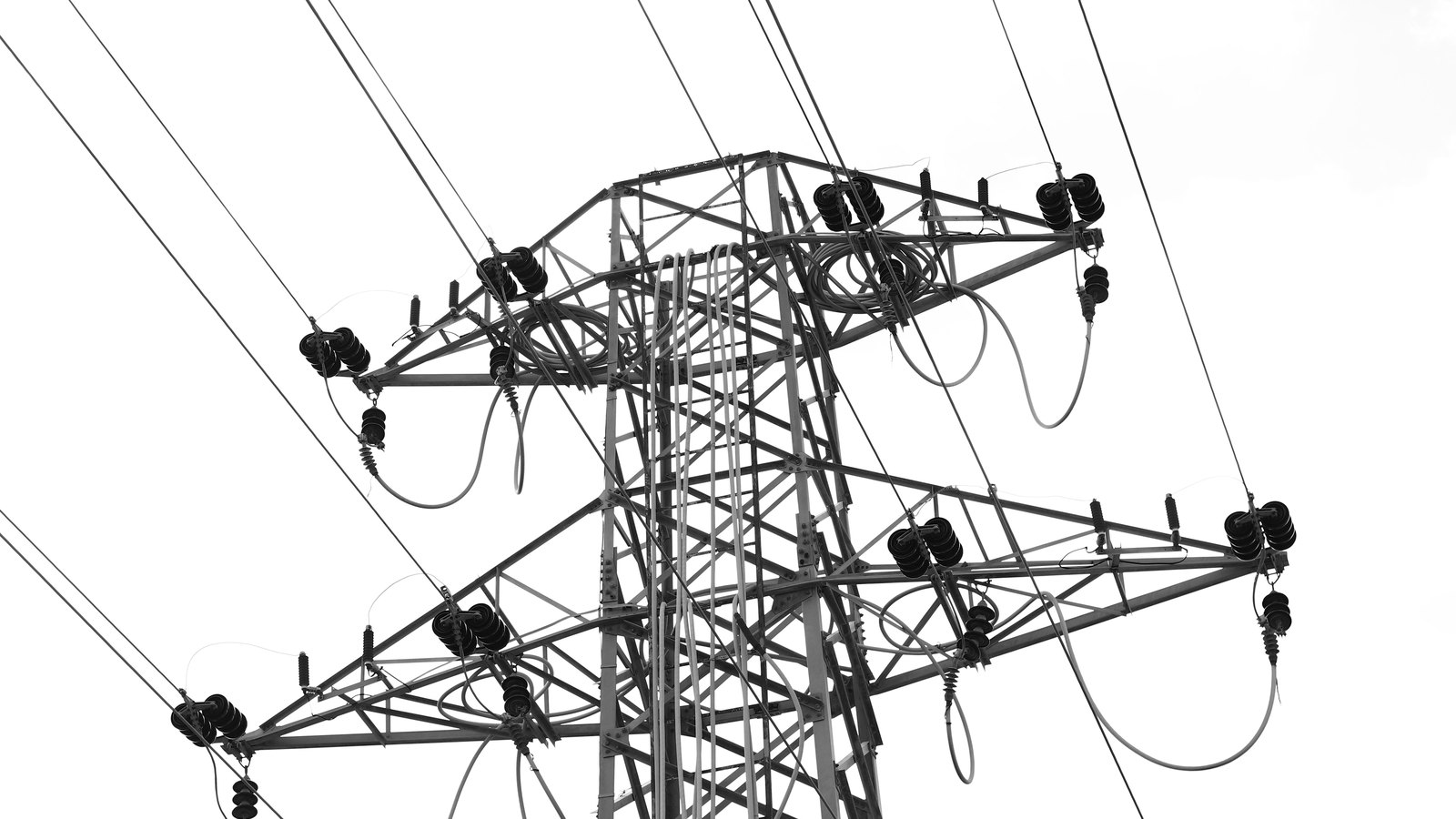 A black and white photograph looking up at the head of a high voltage electricity transmission tower against a blank white sky, its steel lattice arms carrying rows of ribbed insulator stacks with conductors and looping jumper cables running off to both sides, with no ground, horizon or lettering in view.