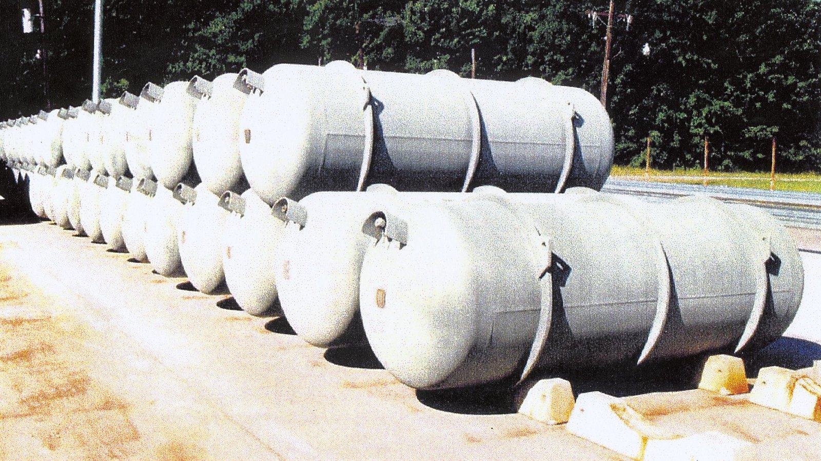 Rows of steel uranium hexafluoride storage cylinders lying on concrete saddles in one of the Department of Energy cylinder storage yards at the Paducah site in Kentucky, with trees behind and a service road to the right.