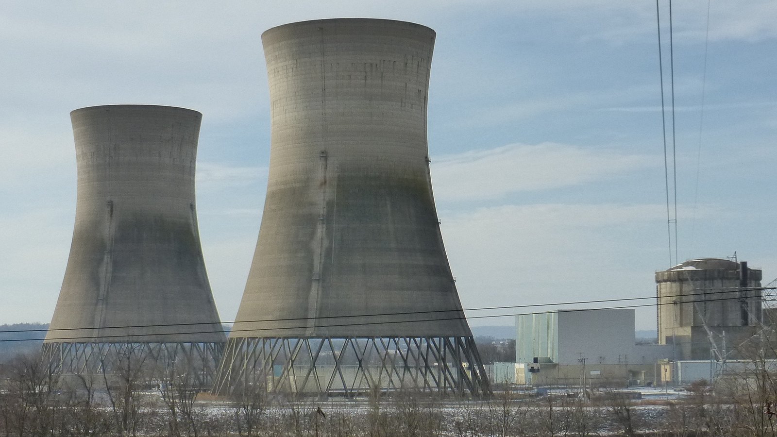 The two dormant cooling towers of Unit 2 at the Three Mile Island Nuclear Generating Station in Pennsylvania, standing empty against a pale winter sky with no vapour rising, the reactor containment building and low plant structures to the right, overhead power lines crossing the frame and snow on the ground.