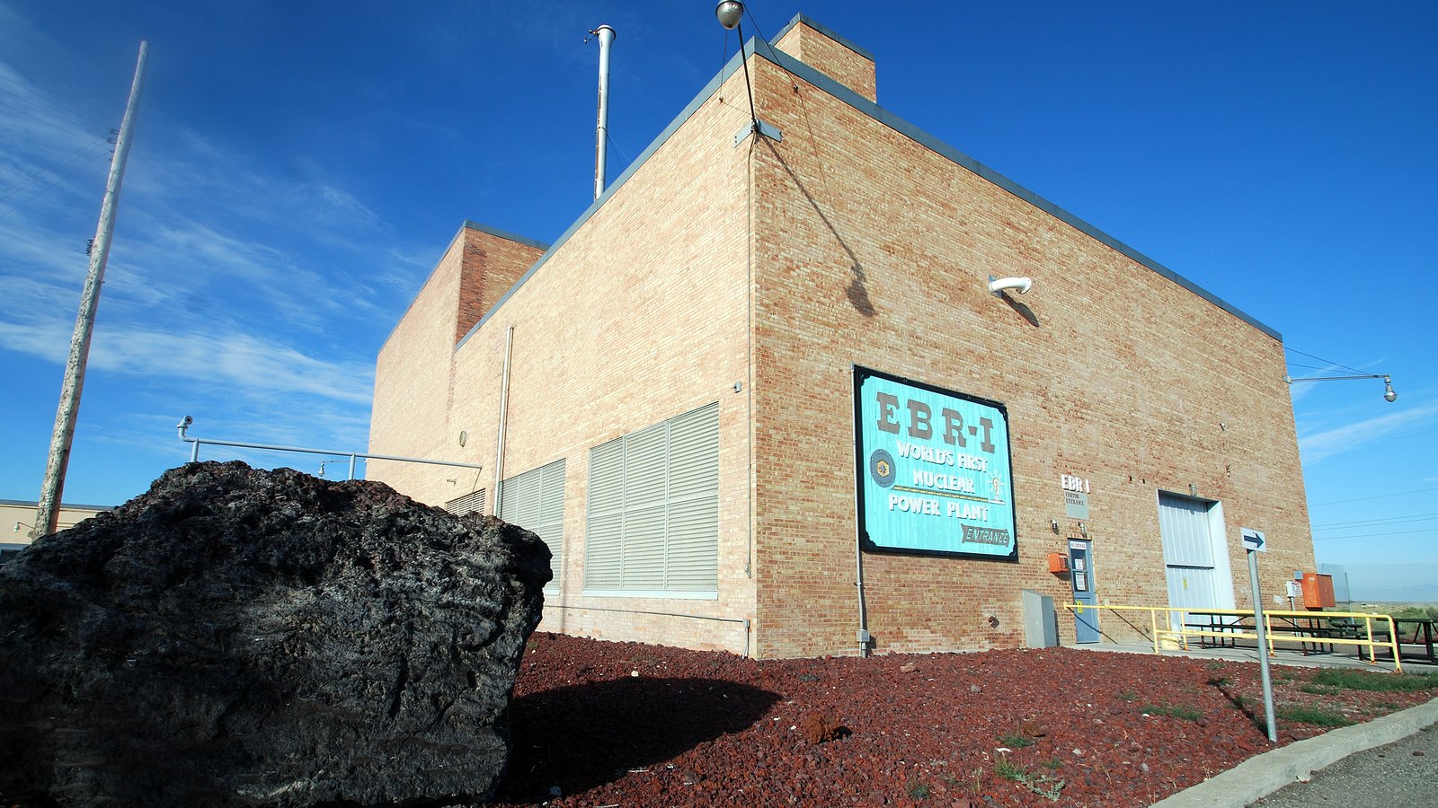 The tan brick Experimental Breeder Reactor I building at Idaho National Laboratory under a blue sky, its own turquoise sign reading EBR-I, World's First Nuclear Power Plant, with a large black lava boulder and red cinder landscaping in the foreground. No people are in view.