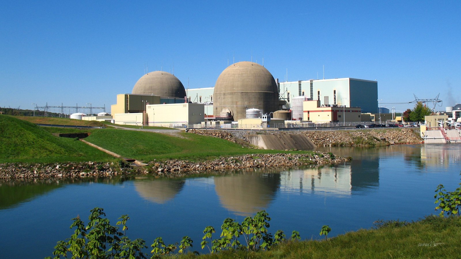 North Anna Power Station in Louisa County, Virginia, seen across the warm water discharge canal: two brown domed reactor containments side by side, with the turbine hall and a pale blue-green service building extending to the right and reflected in the still water, and a grassy bank in the foreground.