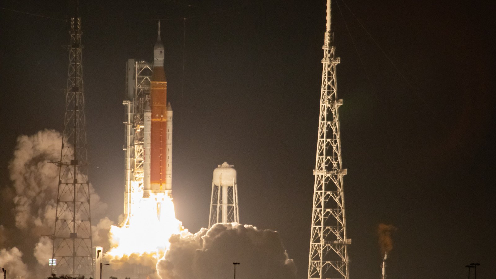 A NASA Space Launch System rocket rising from its pad at night, the exhaust throwing white light across the launch complex, framed between two steel lightning towers with a water tower behind.