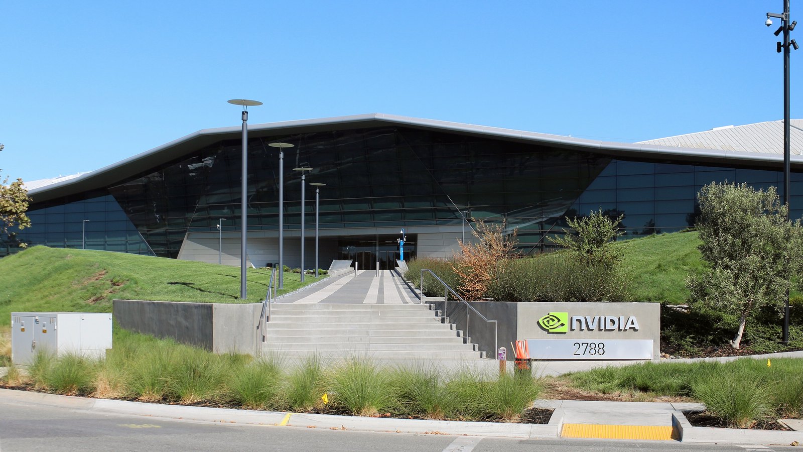 The NVIDIA headquarters building in Santa Clara, California, seen from the road: a long low structure clad in dark tinted glass under a folded silver roofline, set behind a grass bank, with a wide flight of concrete steps rising to a recessed entrance and the company name and eye mark on a low concrete wall beside the steps above the street number 2788, under a clear blue sky.
