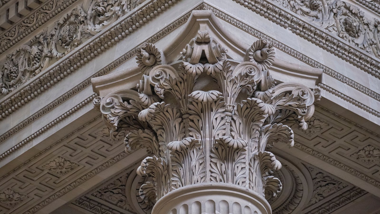 A carved Corinthian column capital of acanthus leaves and scrolls, meeting a stone entablature with egg and dart and Greek key mouldings above it.
