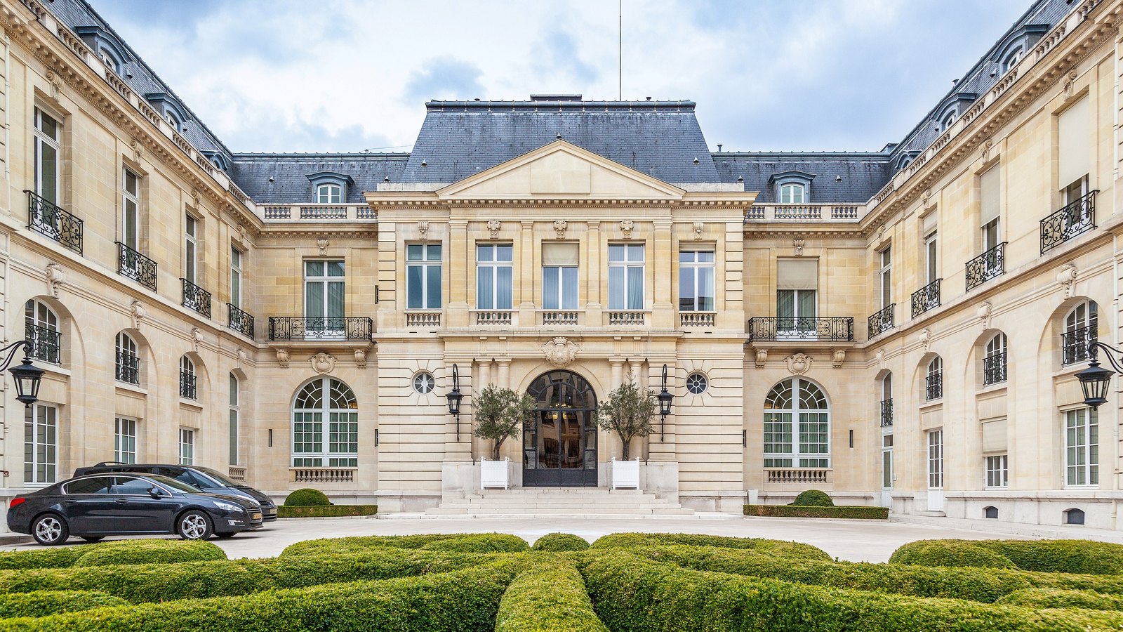 The pale stone Chateau de la Muette in Paris, the headquarters of the OECD, behind clipped hedges beneath a cloudy sky.