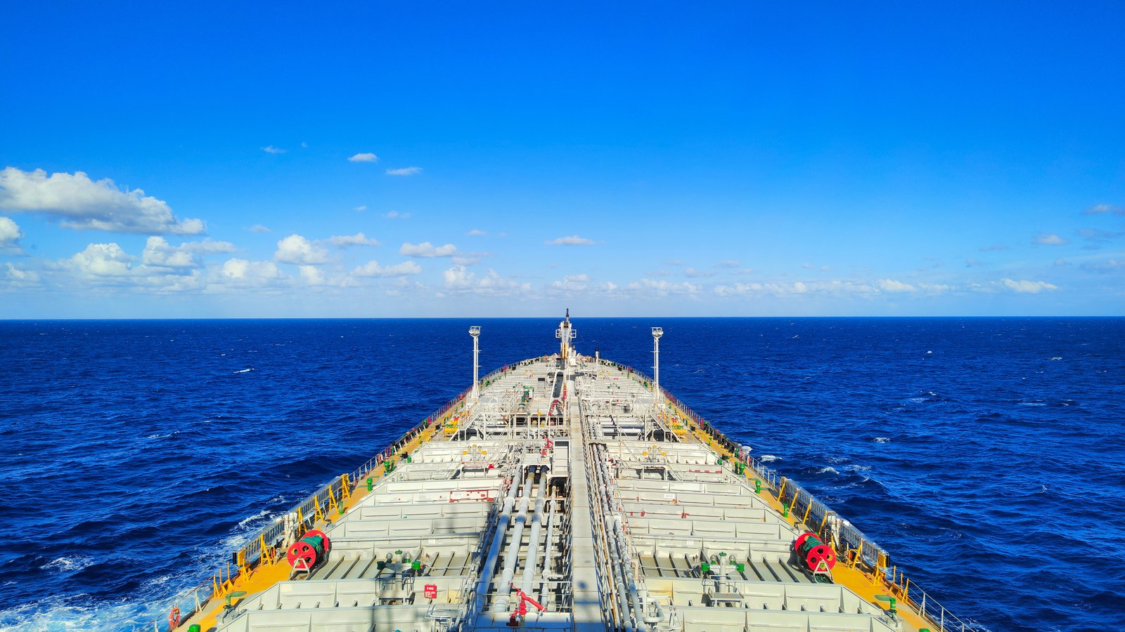 The main deck of a product tanker seen from above the superstructure looking forward over open ocean, twin rows of cargo tank covers and pipework running the length of the deck to the bow, deep blue water on both sides and a line of small cumulus on the horizon. No vessel name, hull number, flag, company mark or legible lettering appears anywhere in the frame. Stock photograph, not a photograph of anything in this story.