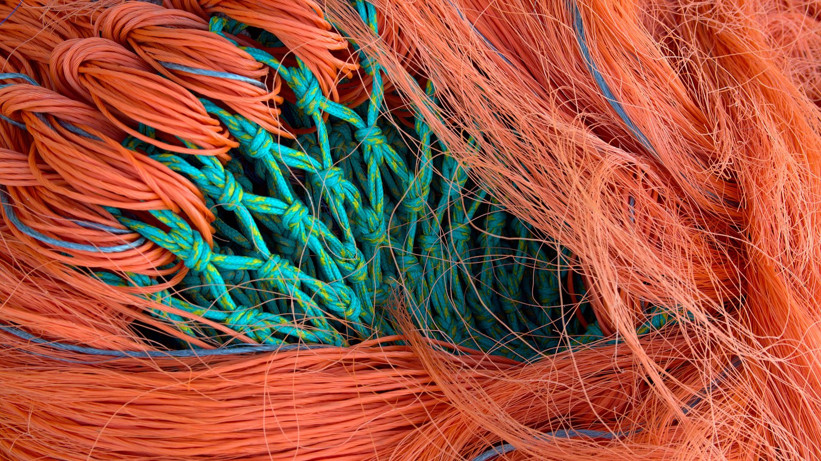 A tight overhead view of commercial fishing net: loose orange monofilament strands sweeping across the frame over a knotted panel of green and yellow mesh beneath, filling the picture edge to edge.