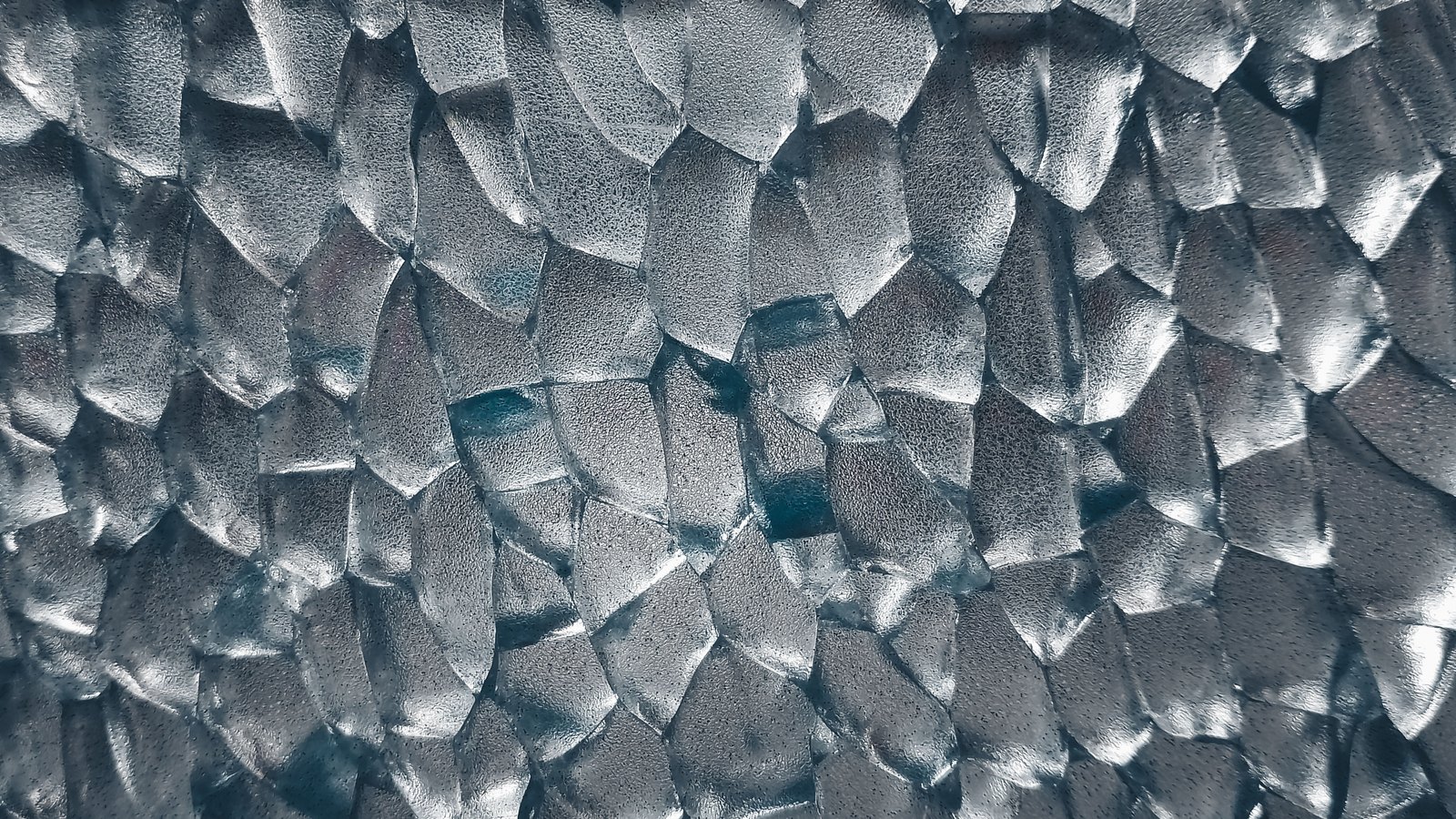 A sheet of textured glass filling the frame, its surface broken into hundreds of overlapping facets in cool silver and pale blue.