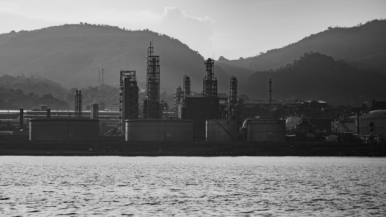 A black and white view across calm water to an oil refinery, with tall distillation columns, cylindrical storage tanks and spherical pressure vessels standing in front of forested hills. No lettering, logo or people are visible.