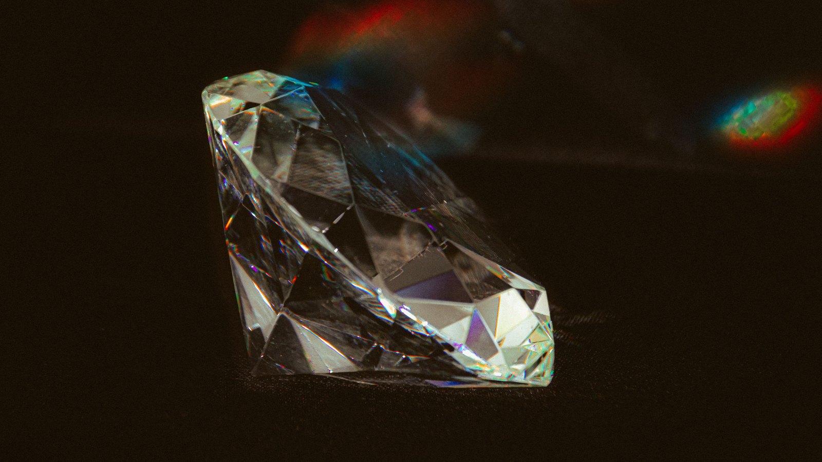 A single brilliant cut stone resting point down on a dark cloth surface, lit from one side so its facets throw small bands of red, green and violet, with the rest of the frame in near darkness.