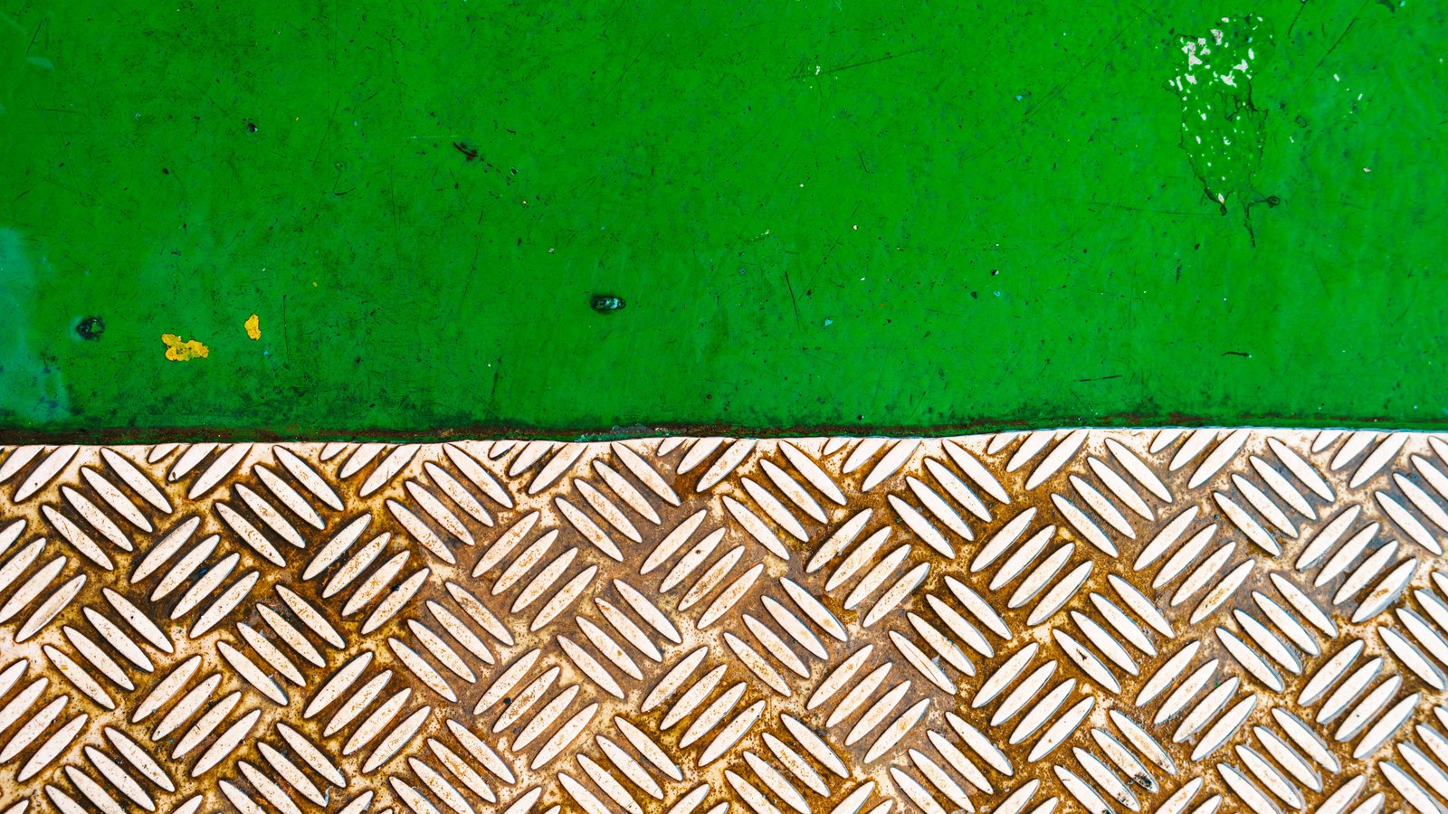 A top-down view of rust-stained steel chequer plate meeting a band of scuffed green deck paint along a straight seam.