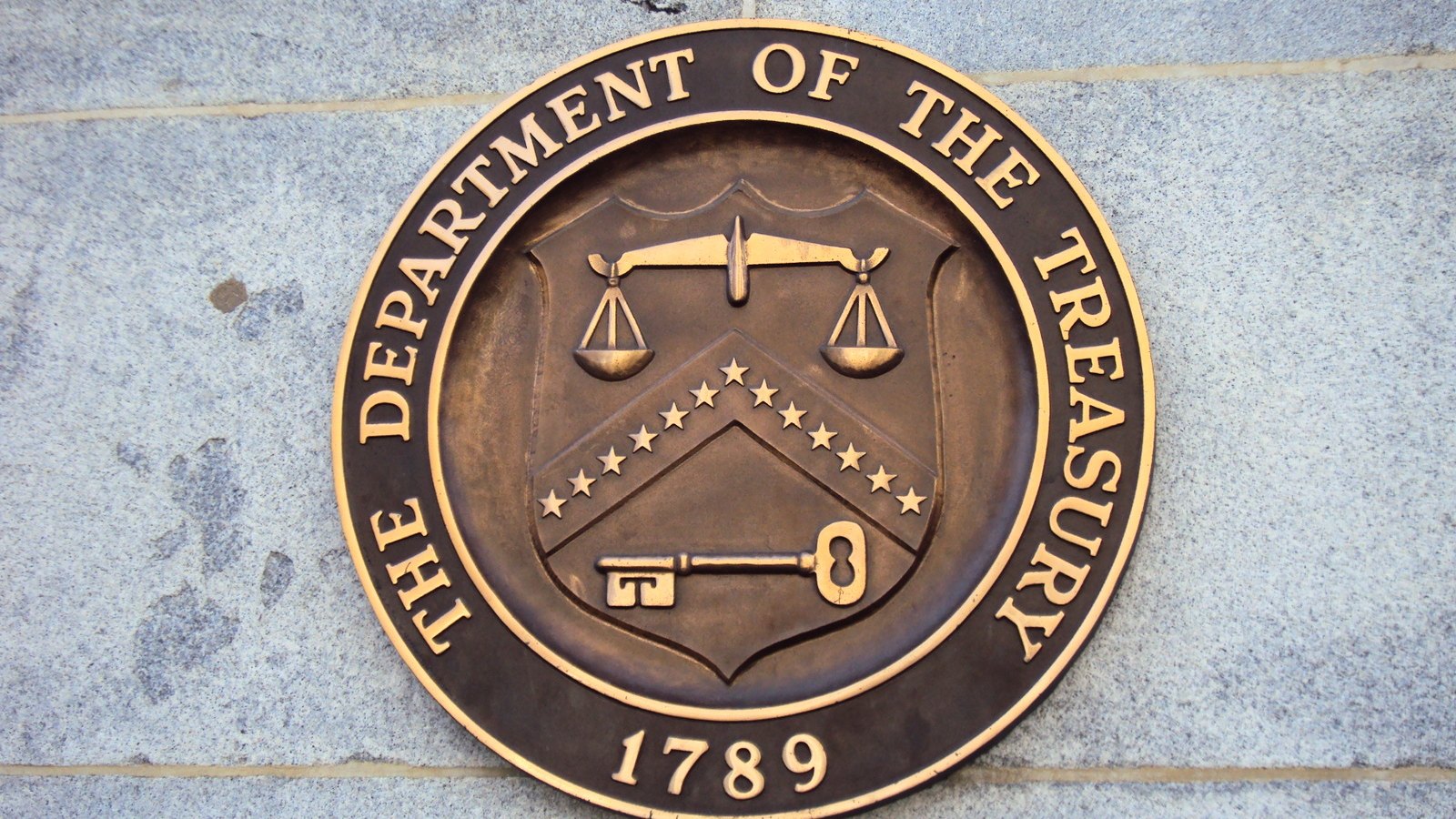 A bronze circular seal fixed to a pale granite wall, its raised outer lettering reading THE DEPARTMENT OF THE TREASURY around a shield bearing scales, a chevron of thirteen stars and a key, with the date 1789 below.