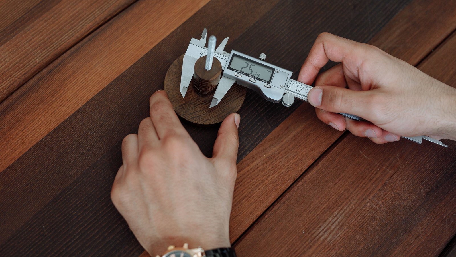 Two hands using a digital caliper to measure a small wooden disc on a dark wooden workbench, the caliper's display showing a reading in millimetres.