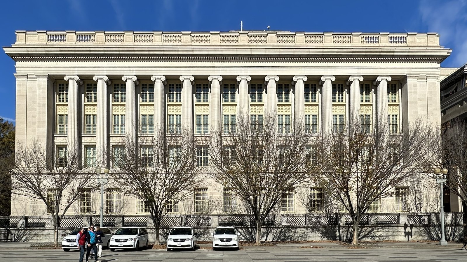 The long colonnaded south front of the Freedman's Bank Building, the Treasury Annex on Pennsylvania Avenue in Washington, seen straight on in winter sunlight above a low iron fence, with bare trees along the pavement and four white vans parked at the kerb. No signage, lettering or logo is legible anywhere in the frame.