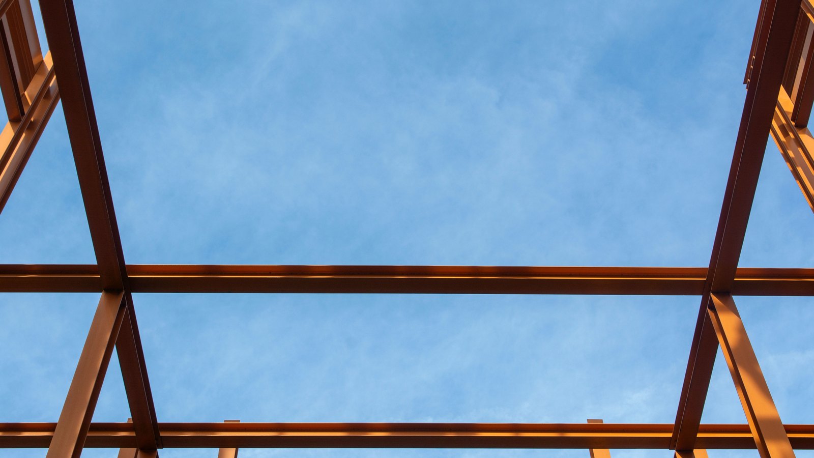 The bare painted steel beams of an unfinished building frame, photographed from directly below against a clear blue sky. Three horizontal girders and several diagonal braces cross the frame. No machinery, lettering or people are visible.