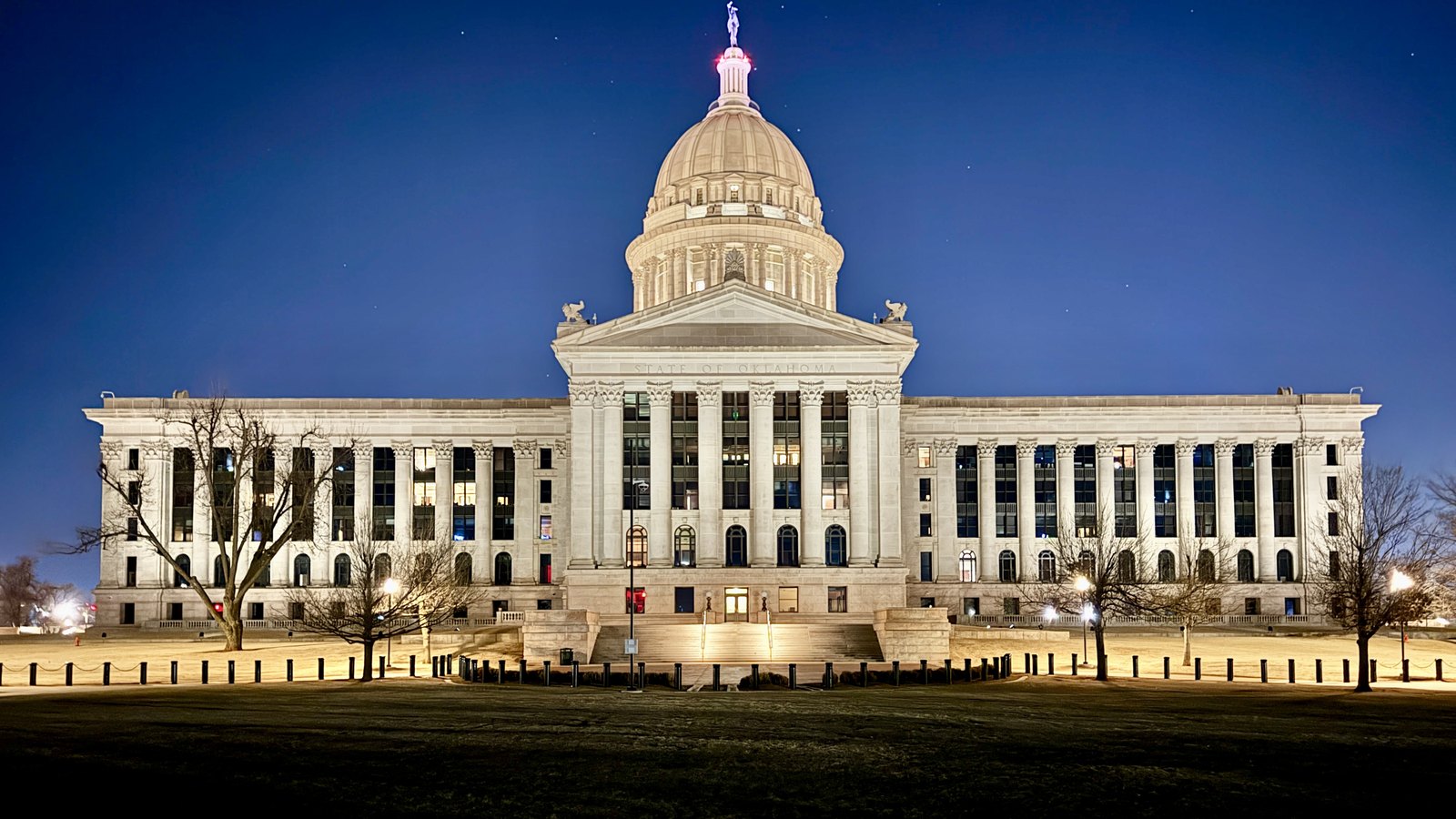 The Oklahoma State Capitol in Oklahoma City floodlit at blue hour, seen straight on across the lawn: a long limestone block with two colonnaded wings, a central portico carrying STATE OF OKLAHOMA carved in the pediment, and the dome above it topped by a bronze figure.