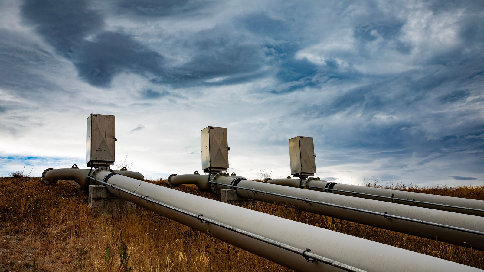 Large parallel pipelines and control housings crossing a grassy field beneath clouds.