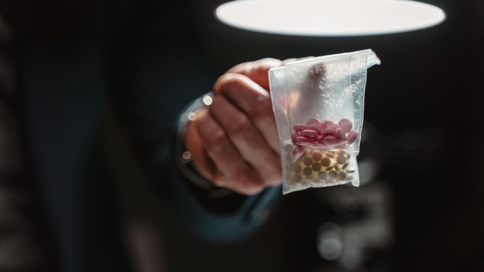 A hand holding a small clear plastic bag of loose pills up toward a bright overhead light in a dark room. Stock photo, not a photograph of this event.