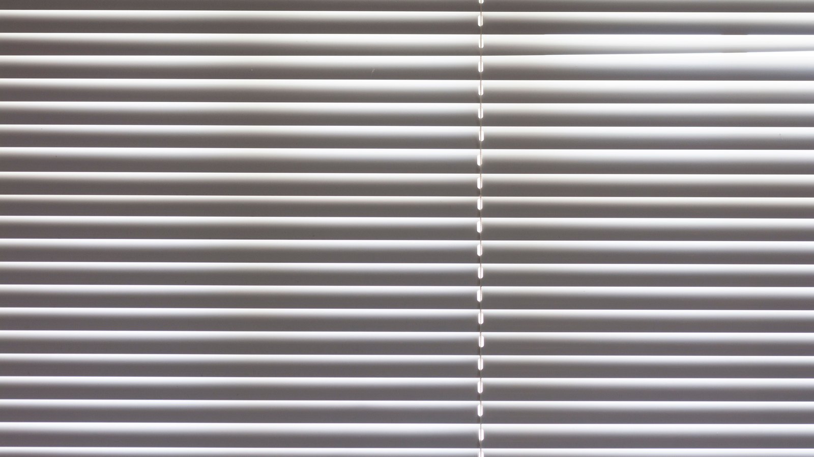 A closed white venetian blind filling the whole frame, its narrow horizontal slats running edge to edge in even rows and lit softly from the left, with one vertical line of small bright gaps down the middle where the lift cords pass through. No window frame, view, text, brand mark or person is in view.