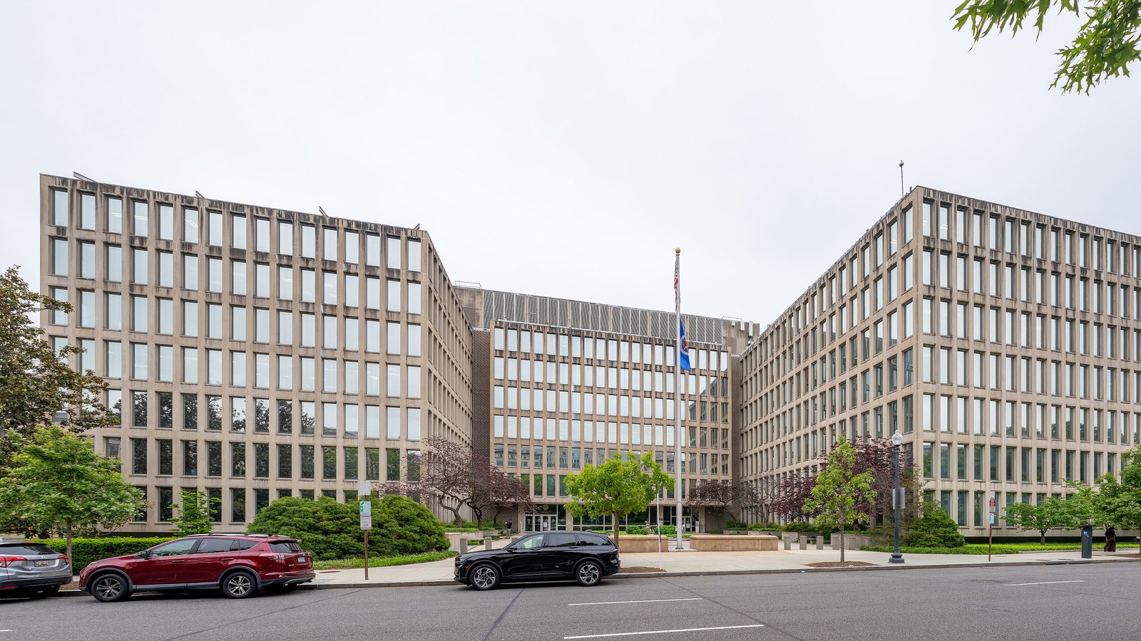 The Theodore Roosevelt Federal Building in Washington, a long pale concrete and glass office block whose two wings meet at a set back centre section, with an American flag on a pole in front of the entrance and cars parked along the kerb under an overcast sky.
