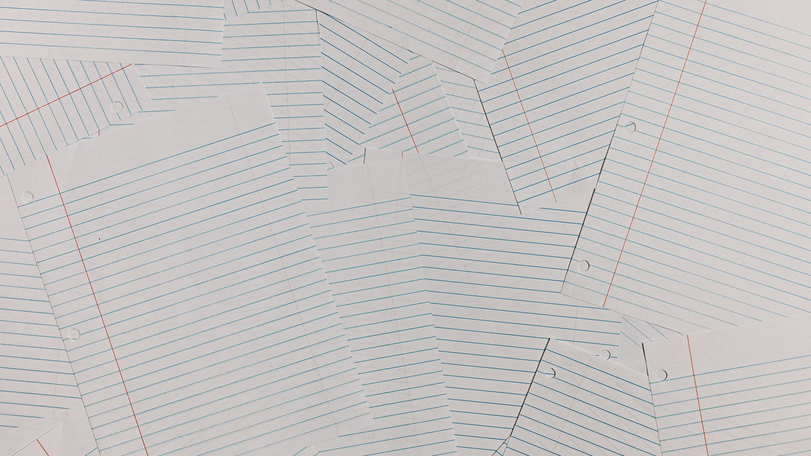 A close photograph of a dozen blank sheets of ruled loose-leaf paper overlapping at angles and filling the frame, pale blue horizontal rules and a red vertical margin on each, with punched holes along some edges. Nothing is written on any sheet and no lettering, object or person is in view.