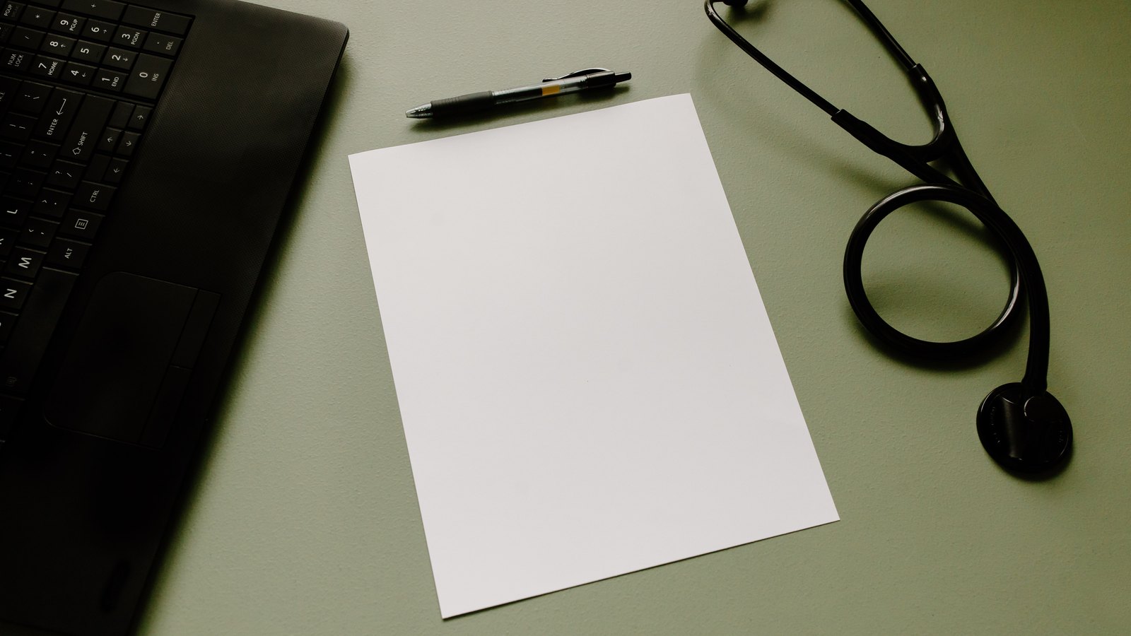 A stethoscope rests beside a blank sheet of paper and a laptop keyboard.