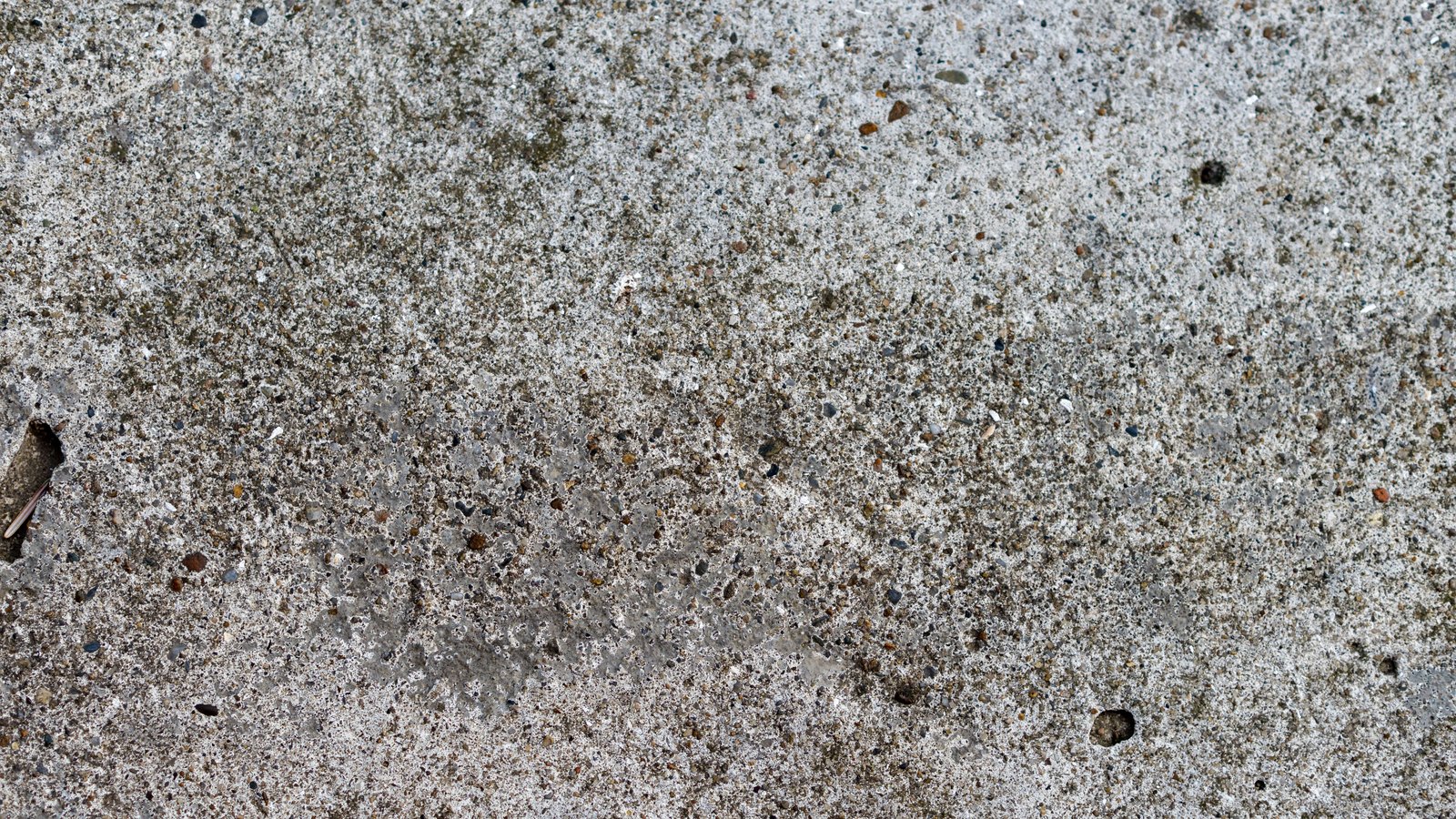 A flat, full-frame macro of a grey concrete surface, its pale cement matrix flecked with small brown, white and dark aggregate stones, with no text, objects or people in view.