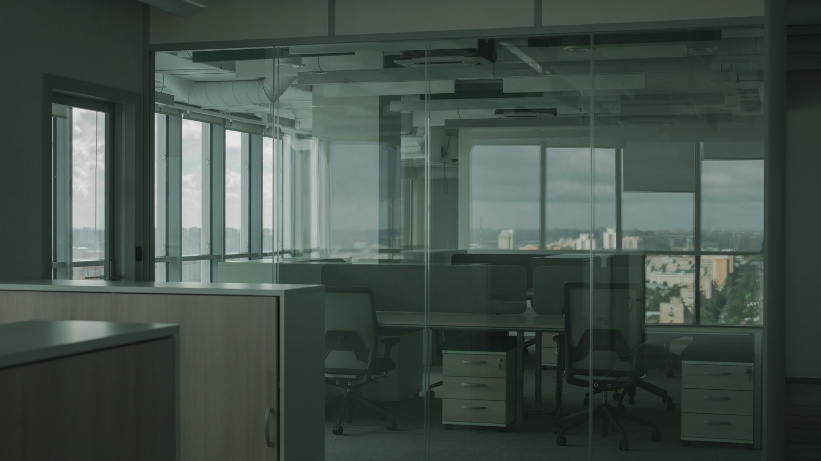 An empty open plan office seen through a glass partition, unoccupied swivel chairs pushed back from two rows of desks with white pedestal units beside them, and a distant grey city skyline through the windows behind. Nobody is present.