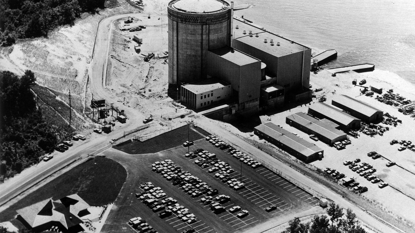 A black and white aerial photograph of the Palisades nuclear plant on the shore of Lake Michigan. A tall cylindrical concrete containment building stands at the centre, with a long flat-roofed turbine hall beside it running out towards the water and low sheds and stacked materials on the open ground to the right. A full car park of parked cars fills the foreground, a wooded bluff runs down the left side, and open water occupies the top right corner. No sign, lettering or person is legible anywhere in the frame.