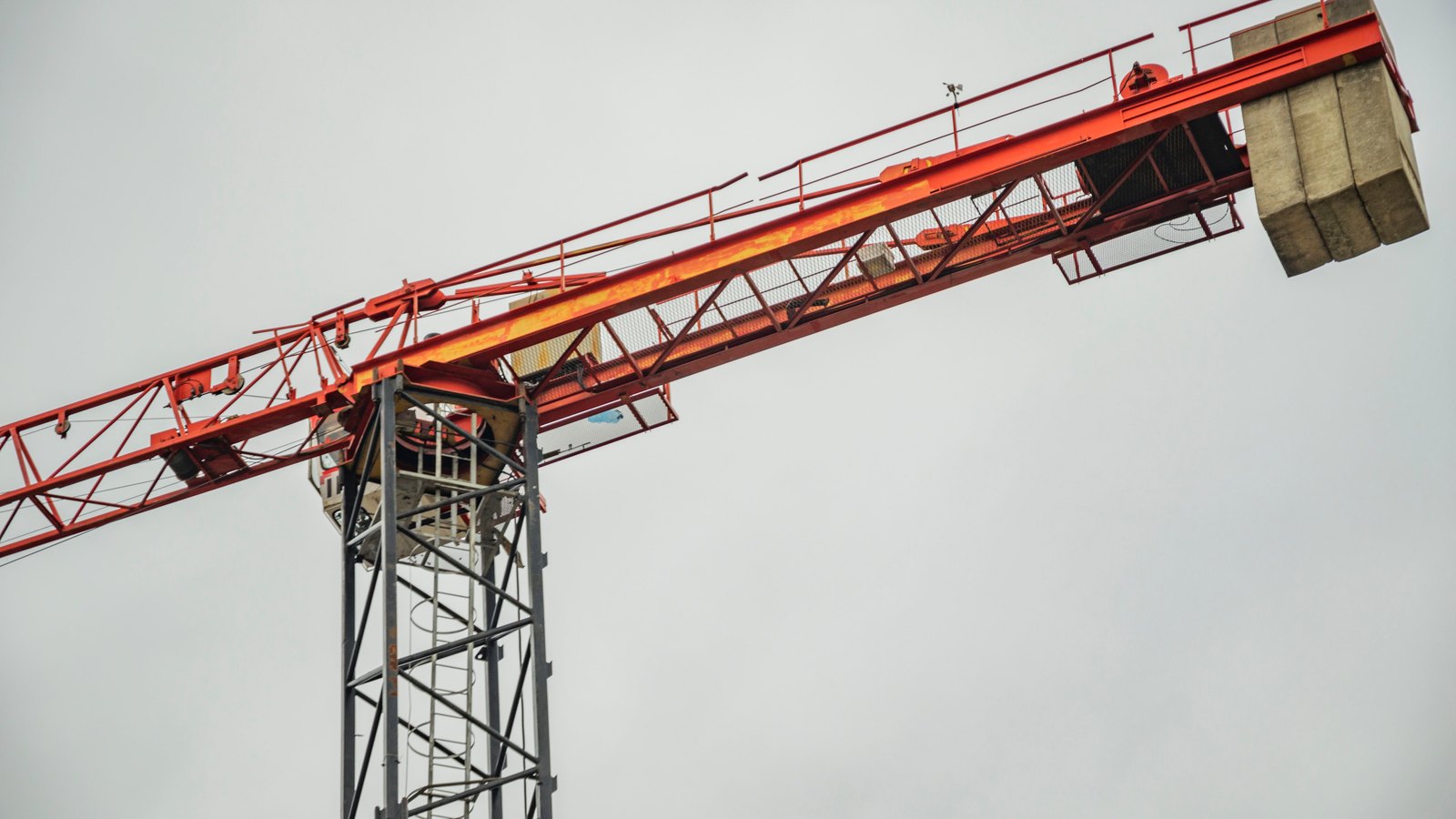 The upper part of a red lattice tower crane seen from below against a plain grey sky, with stacked concrete counterweight blocks on the short arm and a hoist trolley part way along the jib.