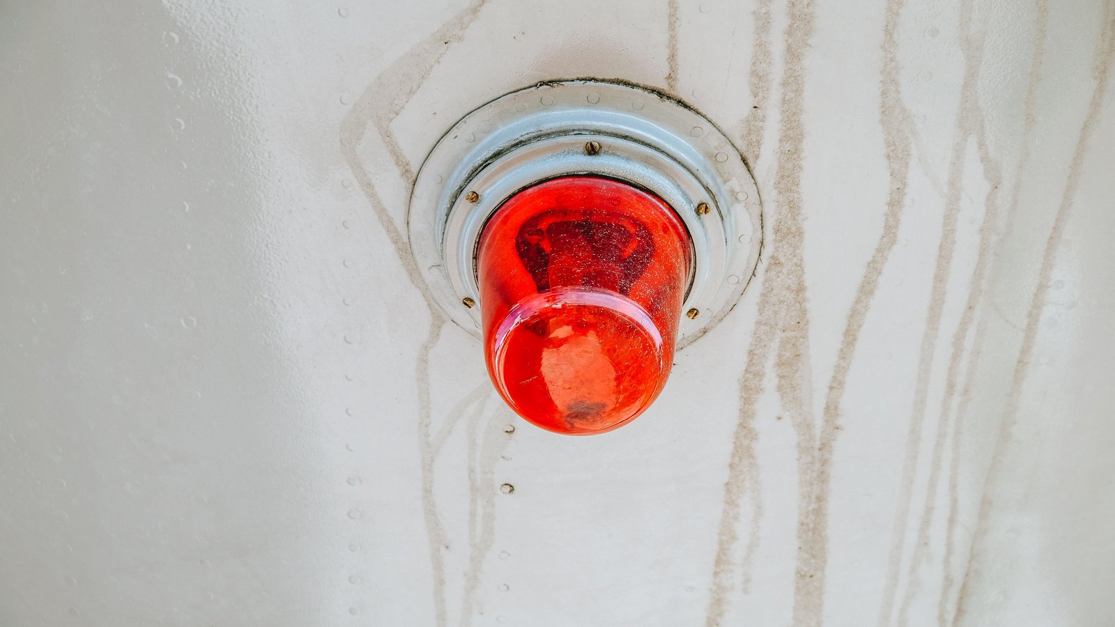 A red emergency beacon lamp set into a weathered white painted metal panel, its domed lens glowing red, with rust streaks running down the paintwork below it. No person, vehicle, road, building, signage or lettering of any kind is visible in the frame. Stock photograph, not a photograph of this event.