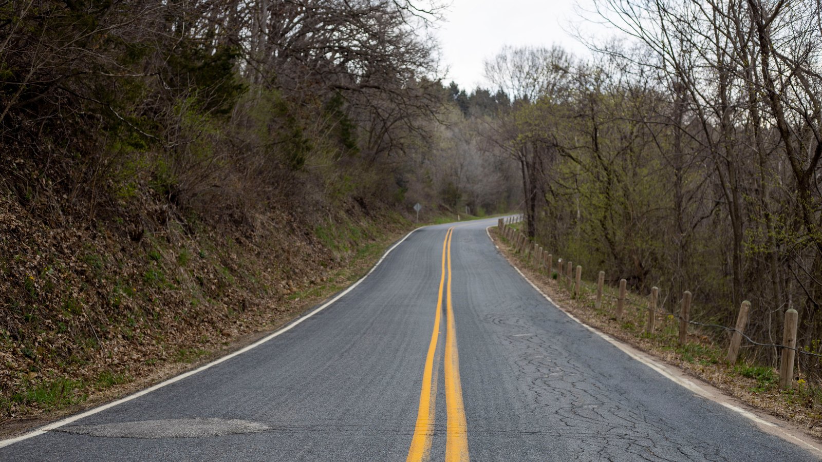 A photograph looking straight down an empty two lane road that runs away from the camera and curves out of sight, with a worn double yellow centre line, white edge lines, a bare wooded bank rising on the left and a low wooden post fence on the right. No vehicles, people, emergency services, readable signage or lettering are visible and no place can be identified from it. A plain road illustration, not a photograph of Palm Beach Boulevard, of Interstate 75, of Fort Myers or of either crash, and not evidence about the condition of any road.