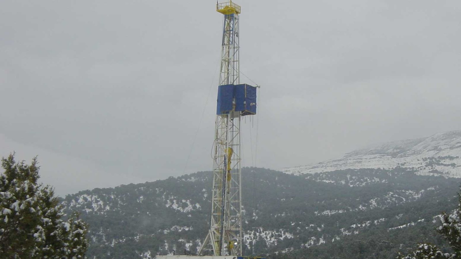 A white and blue drilling rig derrick with a yellow crown platform standing above snow covered pinyon and juniper trees in the Rulison gas field in Garfield County, Colorado, with a snow covered mesa ridge behind it under flat grey cloud.