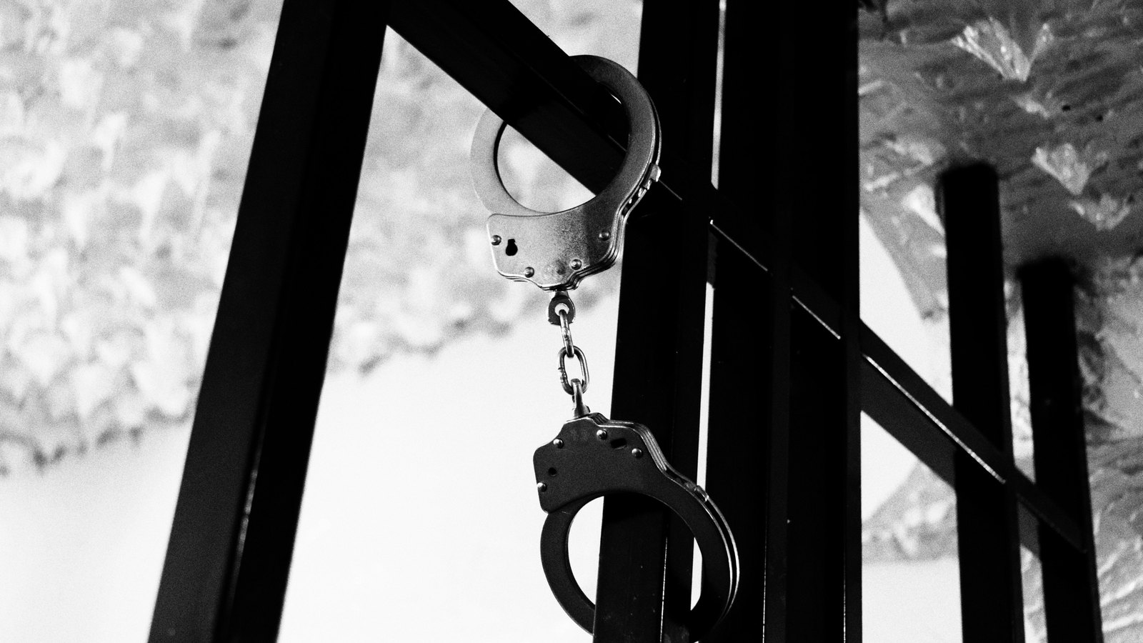 A black and white photograph of a pair of open metal handcuffs hanging from a dark metal railing, one cuff hooked over the bar and the other dangling on its chain against a pale wall. No person, no place, no signage and no lettering appears in the frame. A generic illustration of custody, not a photograph of this event.