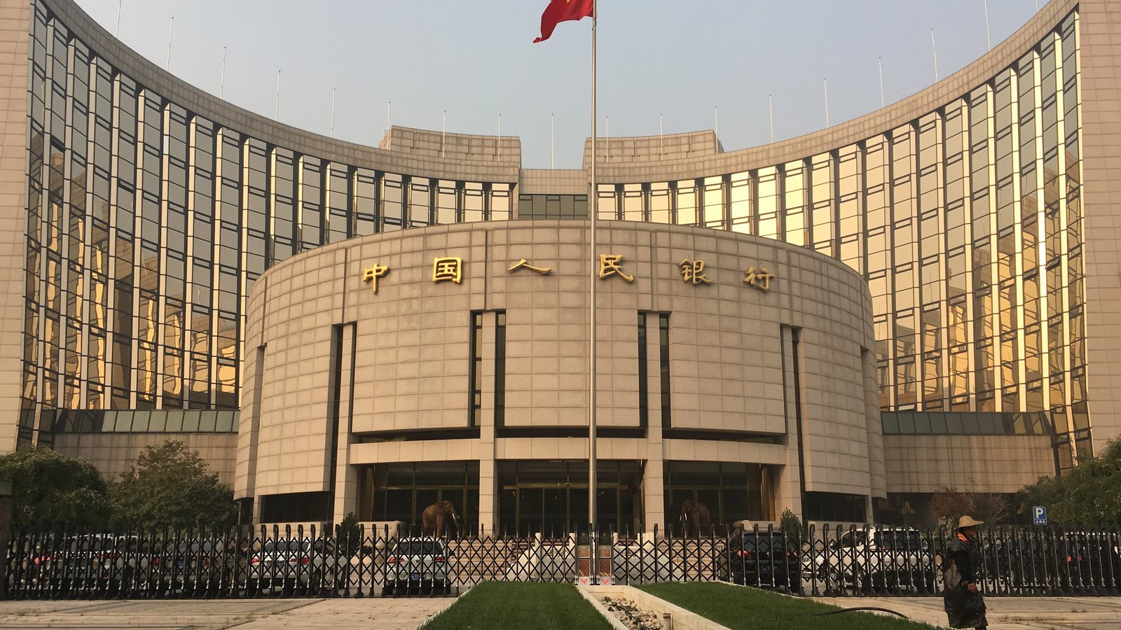 The head office of the People's Bank of China in Beijing under a low sun: a curved glass slab wrapped around a stone drum entrance that carries the bank's name in gold Chinese characters, the national flag on a mast in front of it, and cars parked behind a folding steel gate along the pavement.