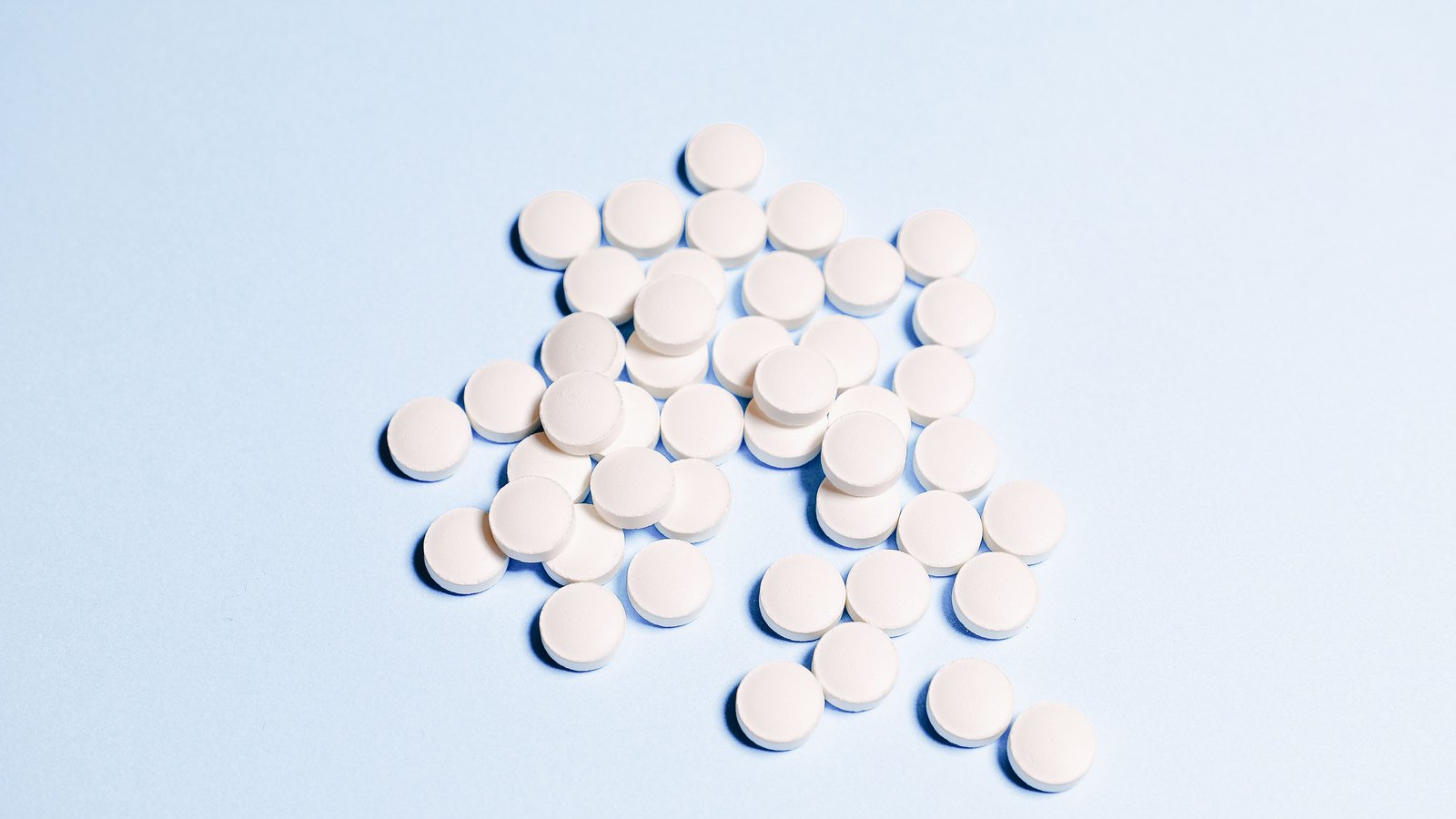 A loose scatter of plain white round tablets, unmarked and uncoated, on a pale blue seamless background.