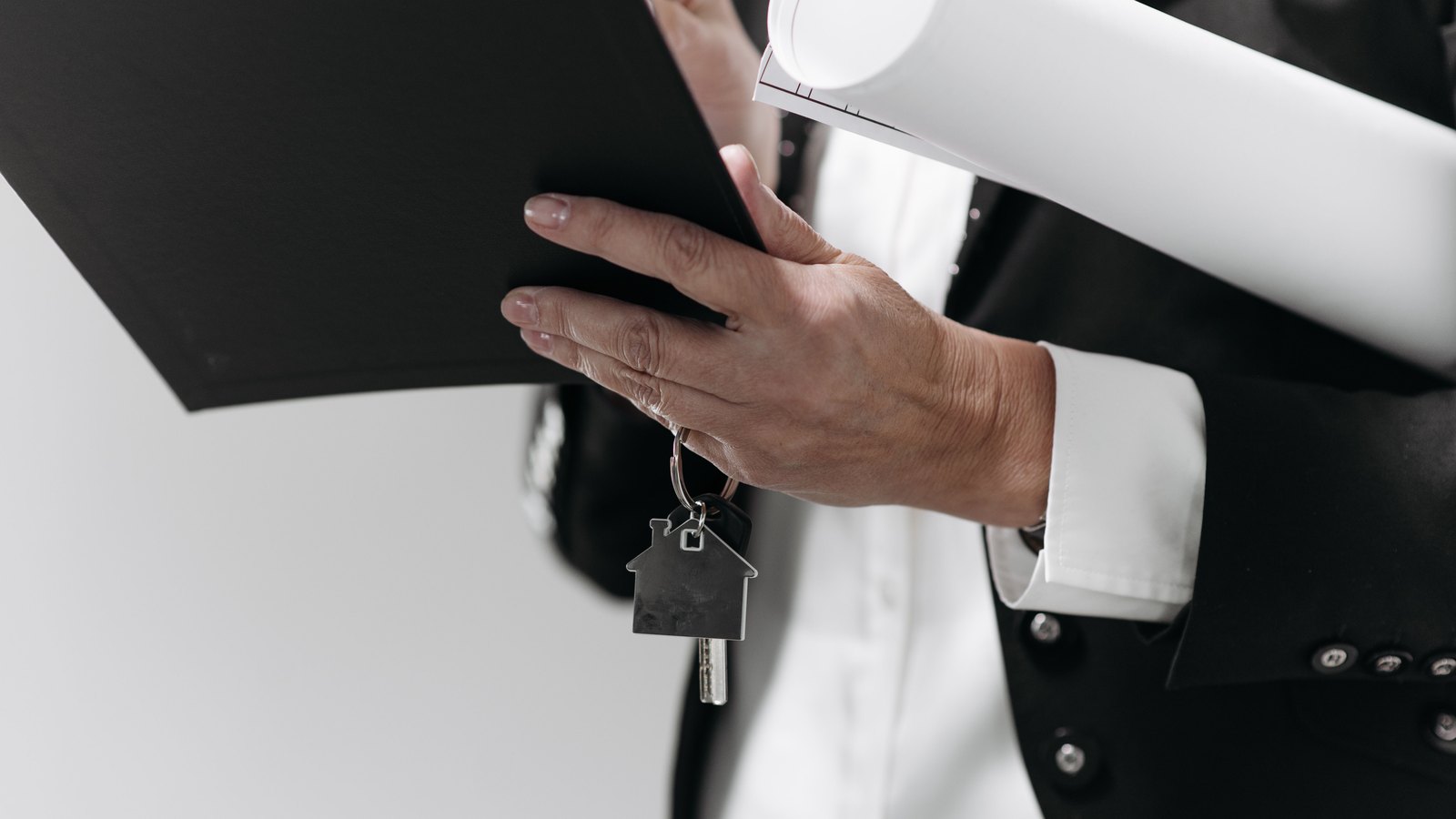 A hand holds a keyring with a house-shaped fob beside an unmarked folder and rolled plans.