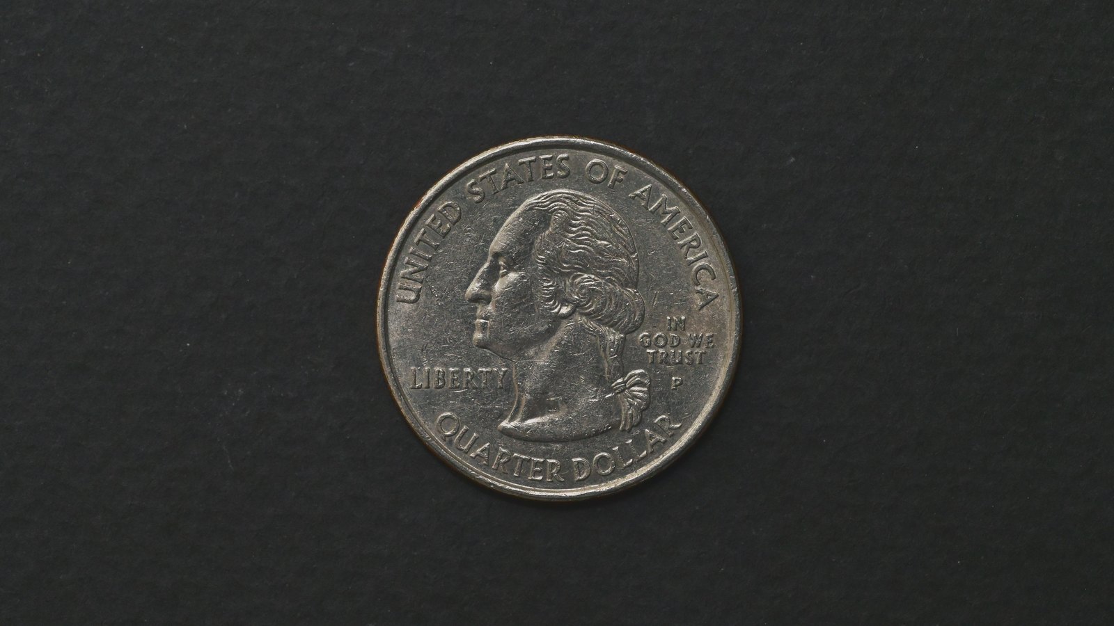 A single circulated United States quarter dollar lying face up on a plain dark grey surface, filling the middle of the frame. The obverse legend reads UNITED STATES OF AMERICA above the profile of George Washington, with LIBERTY to the left, IN GOD WE TRUST and a P mint mark to the right, and QUARTER DOLLAR curving along the bottom.